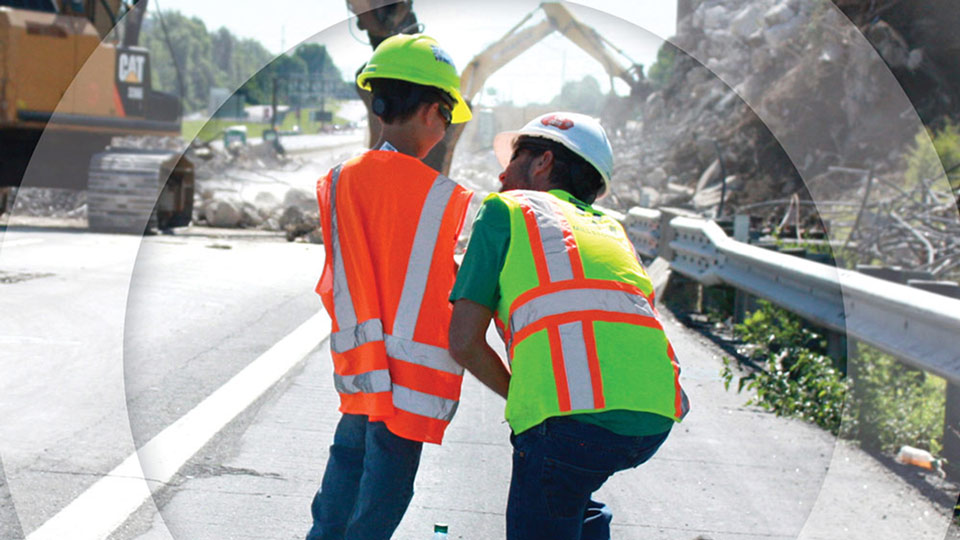 Father and son wearing high-visibility clothing and hard hats standing outside a highway construction zone.