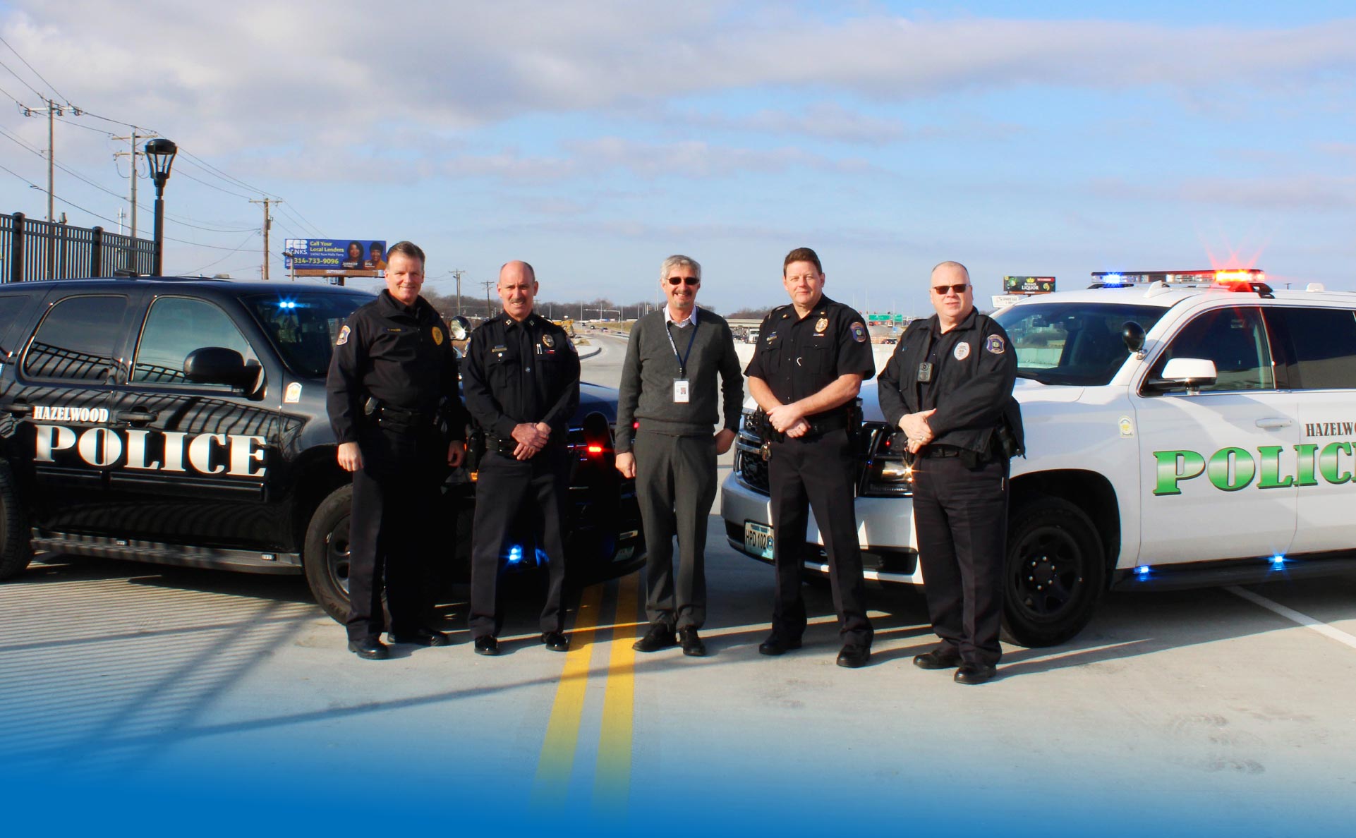 Four police officers and a community leader standing on a newly built bridge in front of two police sport utility vehicles with the words Hazelwood Police along the sides.